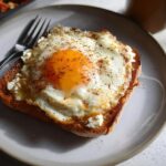 A close-up of a crispy feta fried egg served on a slice of toasted bread, seasoned with herbs.