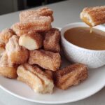 A pile of fluffy air fryer churro bites coated in cinnamon sugar, with one bite being dipped into a bowl of caramel sauce.