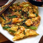 A close-up of a freshly cooked Korean pancake (Pajeon) topped with shrimp and green onions, served on a white plate with chopsticks.