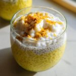 A close-up of a glass filled with vibrant yellow Lemon Cream Chia Pudding, topped with creamy white coconut and a sprinkle of zest.