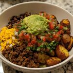 A delicious Loaded Potato Taco Bowl filled with seasoned ground beef, black beans, shredded cheese, pico de gallo, guacamole, and roasted potatoes.