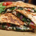 Close-up of Mediterranean Quesadillas filled with spinach, feta, mozzarella, and red onion, served with a side of fresh spinach and cherry tomatoes.