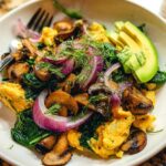 A vibrant bowl of Mushroom Spinach Scrambled Eggs topped with red onion, fresh dill, and sliced avocado.