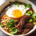 A bowl of Slow Cooker Beef Ramen Noodles topped with a perfectly fried egg, shredded beef, and fresh cilantro.