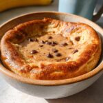A close-up of a baked protein pancake bowl topped with creamy sauce and chocolate chips.