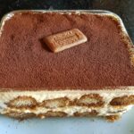 A square Biscoff Tiramisu in a glass dish, topped with cocoa powder and a Biscoff cookie.