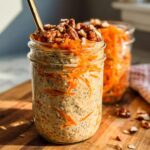 A close-up of Carrot Cake Overnight Oats in a mason jar, topped with shredded carrots and pecans, with another jar in the background.
