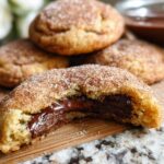 A close-up of a chewy churro cookie, broken in half to reveal a rich chocolate filling. The cookie is coated in cinnamon sugar.