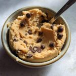 A close-up shot of delicious protein cookie dough with chocolate chips in a bowl, ready to be eaten with a spoon.