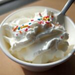 A bowl of creamy frozen Greek yogurt topped with whipped cream and colorful sprinkles, with a spoon.