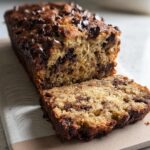 A close-up of a High Protein Chocolate Banana Bread loaf with a slice cut, showing chocolate chips throughout.