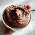 A close-up of a bowl of rich, dark chocolate pudding, topped with chocolate shavings, with a spoon resting in it. This is a High Protein Chocolate Pudding.