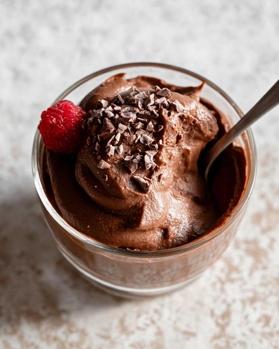 A close-up of a glass bowl filled with decadent High Protein Chocolate Pudding, topped with chocolate shavings and a fresh raspberry.