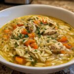 A close-up of a bowl of Lemon Chicken Orzo Soup With Fresh Herbs, featuring tender chicken, orzo pasta, carrots, and parsley.