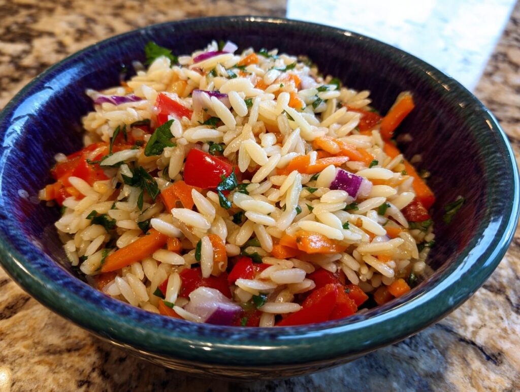 A close-up of a bowl filled with lemony orzo salad, featuring orzo pasta, diced tomatoes, carrots, red onion, and parsley.