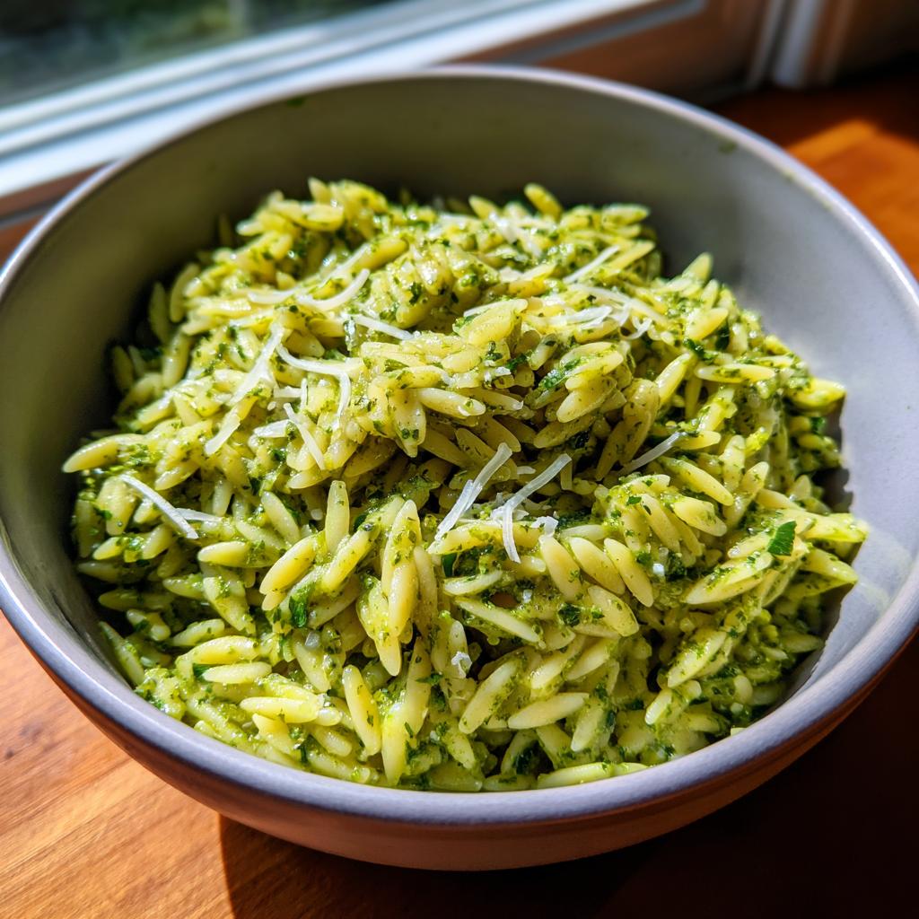 A close-up shot of a bowl filled with bright green pesto orzo pasta, topped with shredded Parmesan cheese.