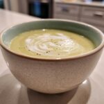 A close-up of a bowl of rich and silky potato leek soup, swirled with cream and garnished with pepper.